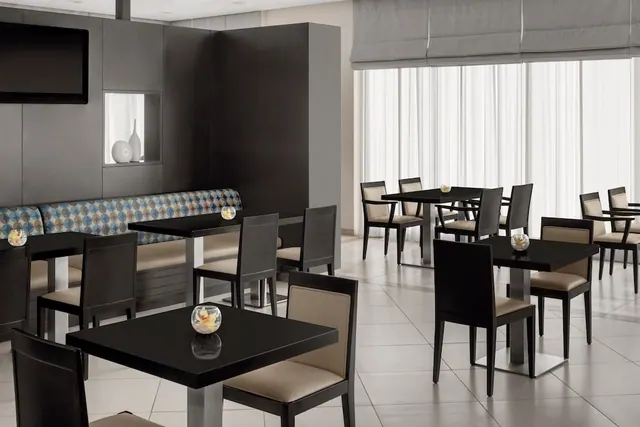 A restaurant dining room with black tables, beige upholstered chairs, a patterned banquette along the wall, and a wall-mounted television beside large windows covered with sheer curtains.