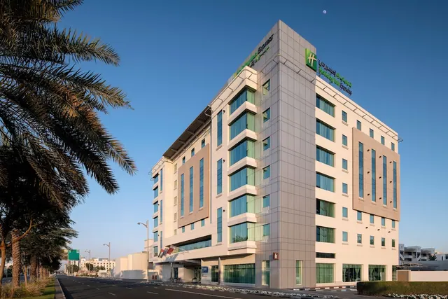 The exterior of a multi-storey Holiday Inn Express hotel with glazed facades, palm trees lining the street, and a clear blue sky.