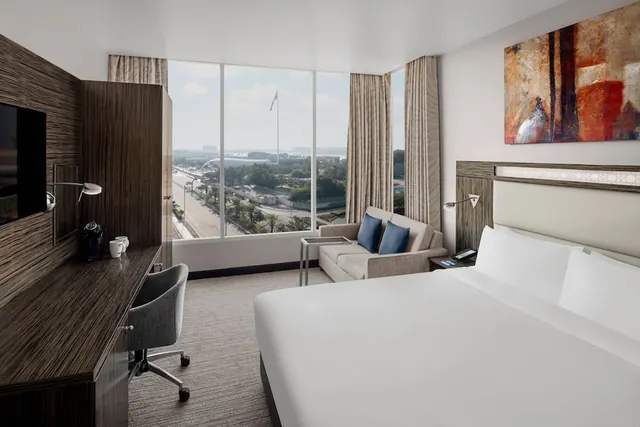 A hotel bedroom with a king bed, a grey sofa with blue cushions, a wooden desk with a chair, and floor-to-ceiling windows looking onto a road lined with palm trees.