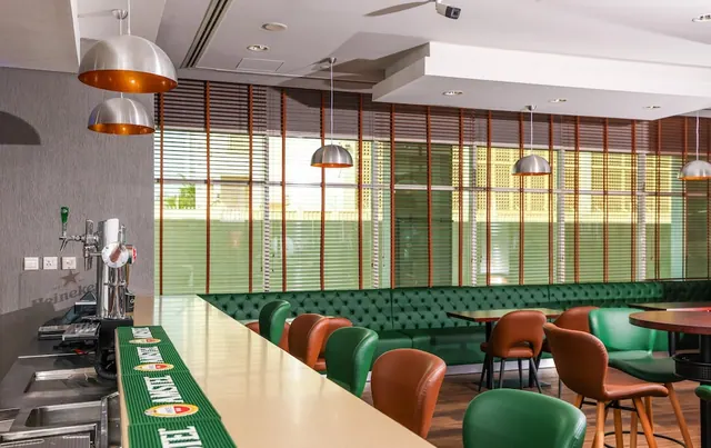 A bar counter with draught taps, green tufted banquette seating, leather chairs and pendant lamps beside a window covered by blinds.