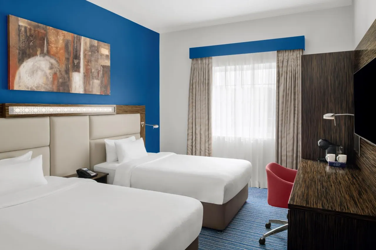 A twin bedroom with two white beds, a blue accent wall with a painting, a work desk with a red chair, a wall-mounted television and a kettle on the desk.