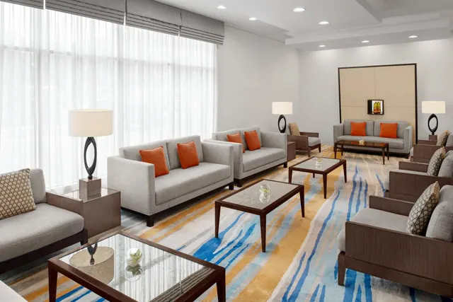 A lounge seating area with grey sofas, orange cushions, glass-topped coffee tables and a patterned rug beside tall sheer-curtained windows.