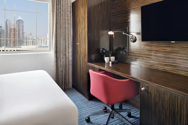 A hotel bedroom with a white bed, a wood-panelled desk with a red chair and wall-mounted television, and a window showing a city skyline with tall towers.