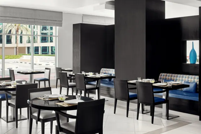 A restaurant dining room with black tables and chairs, blue banquette seating, and large windows showing a palm tree and buildings outside.