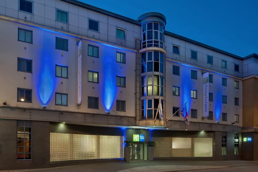 Exterior of a Holiday Inn Express building at dusk, lit with blue uplighting, with the glazed entrance at street level.