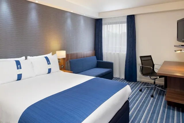 A double bed with blue and white bedding sits beside a blue sofa, a desk with a chair, and a wall-mounted television in a hotel room.