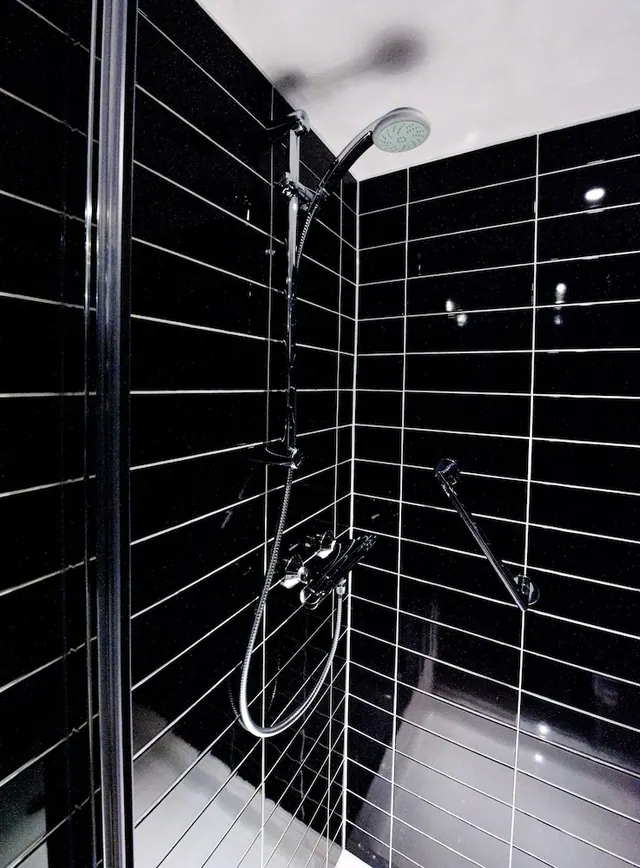 A walk-in shower with black wall tiles, a fixed overhead showerhead, a handheld shower on a rail, and a glass screen on the left.