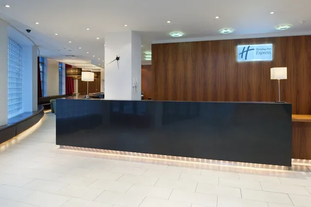 A reception desk with a dark front stands in a lobby beneath a lit Holiday Inn Express sign on a wood-panelled wall, with a curved bench seating area and a glass block wall to the left.