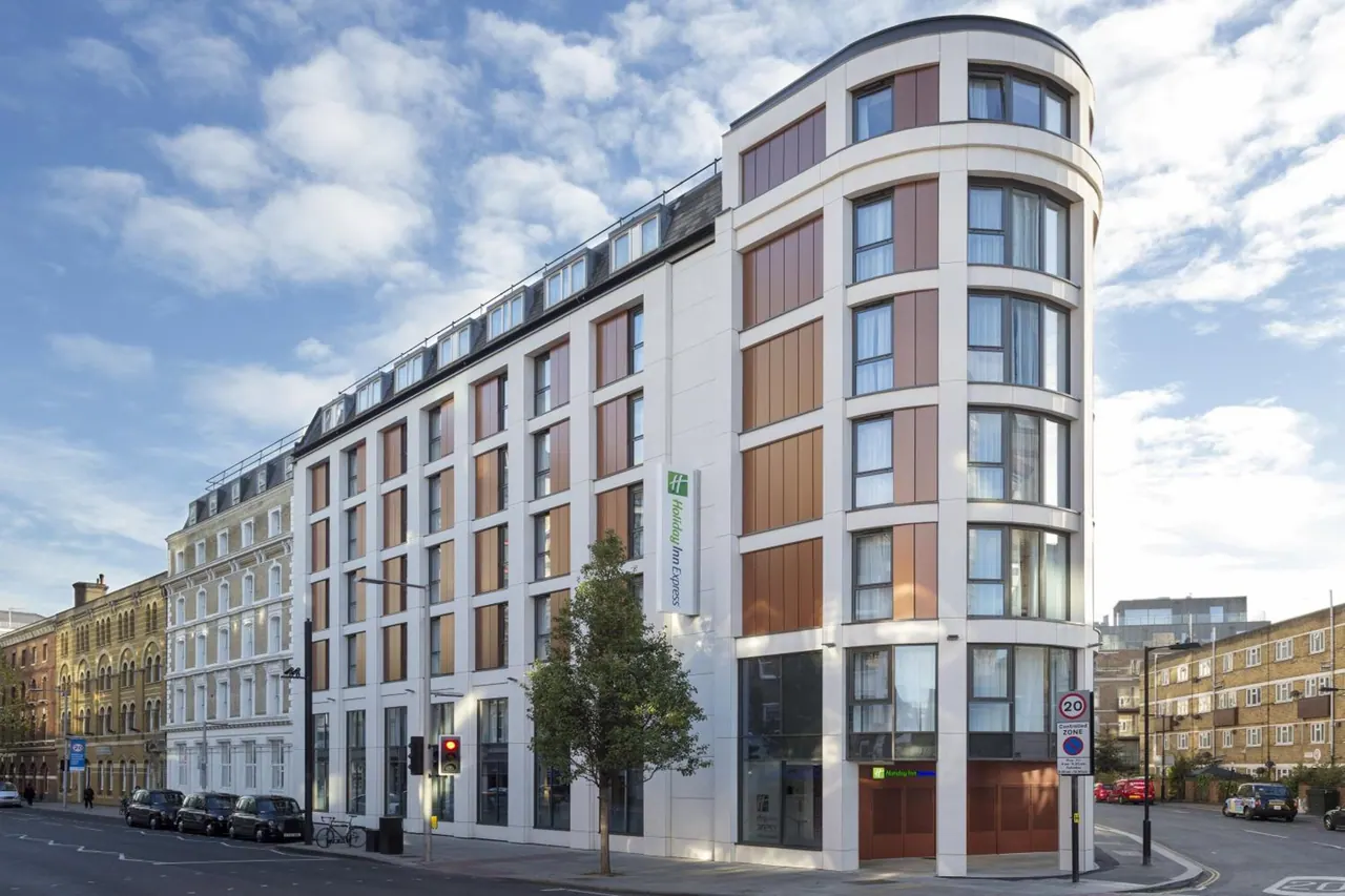 Exterior of a white curved Holiday Inn Express building with bronze panels on a London street, with parked black cabs and a tree at the corner.
