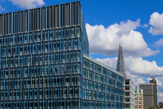 A modern blue glass office building facade stands in front of a blue sky with clouds, with The Shard spire visible behind it.