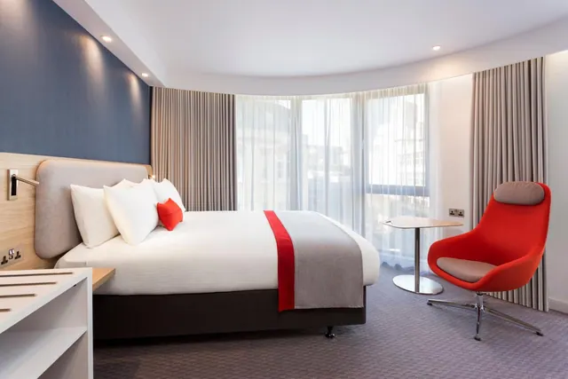 A bright hotel bedroom with a king bed with red accent cushions and throw, a red armchair beside a small round table, and tall windows with sheer curtains.