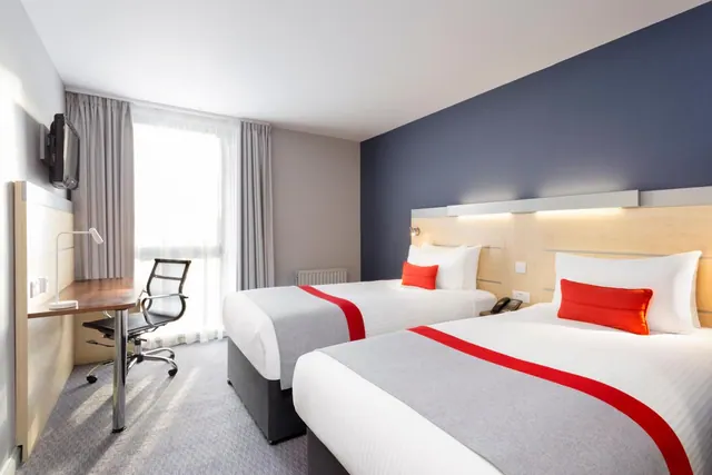 A hotel room with two single beds with red and grey throws, a desk with a chair, and a wall-mounted television.