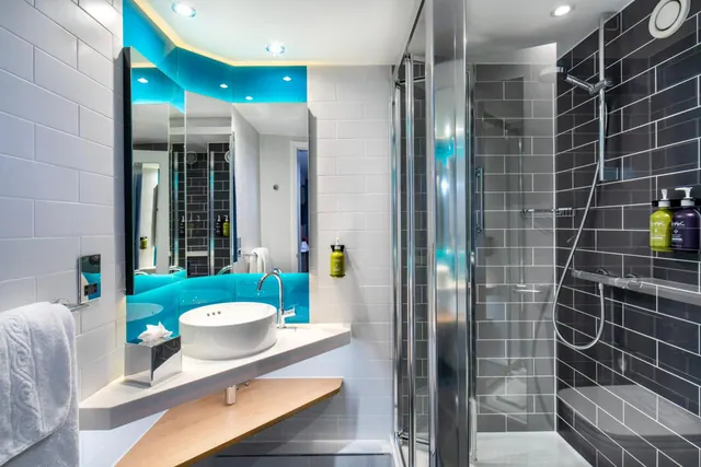 A modern bathroom with a round basin on a floating vanity, a wall mirror with turquoise trim, and a walk-in glass shower with a grab rail.