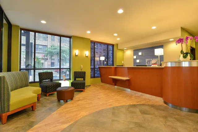 The lobby has a reception desk on the right, green upholstered seating and a round brown ottoman, with large windows looking out onto a city street.