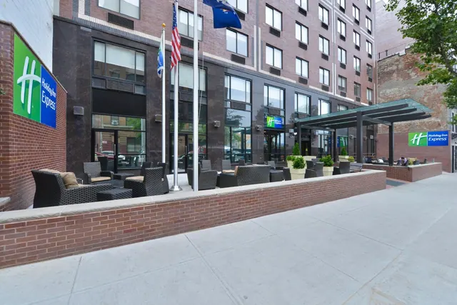 Exterior of a Holiday Inn Express building with an outdoor seating area of wicker chairs and low tables, flags on poles, and a canopy over the sidewalk.