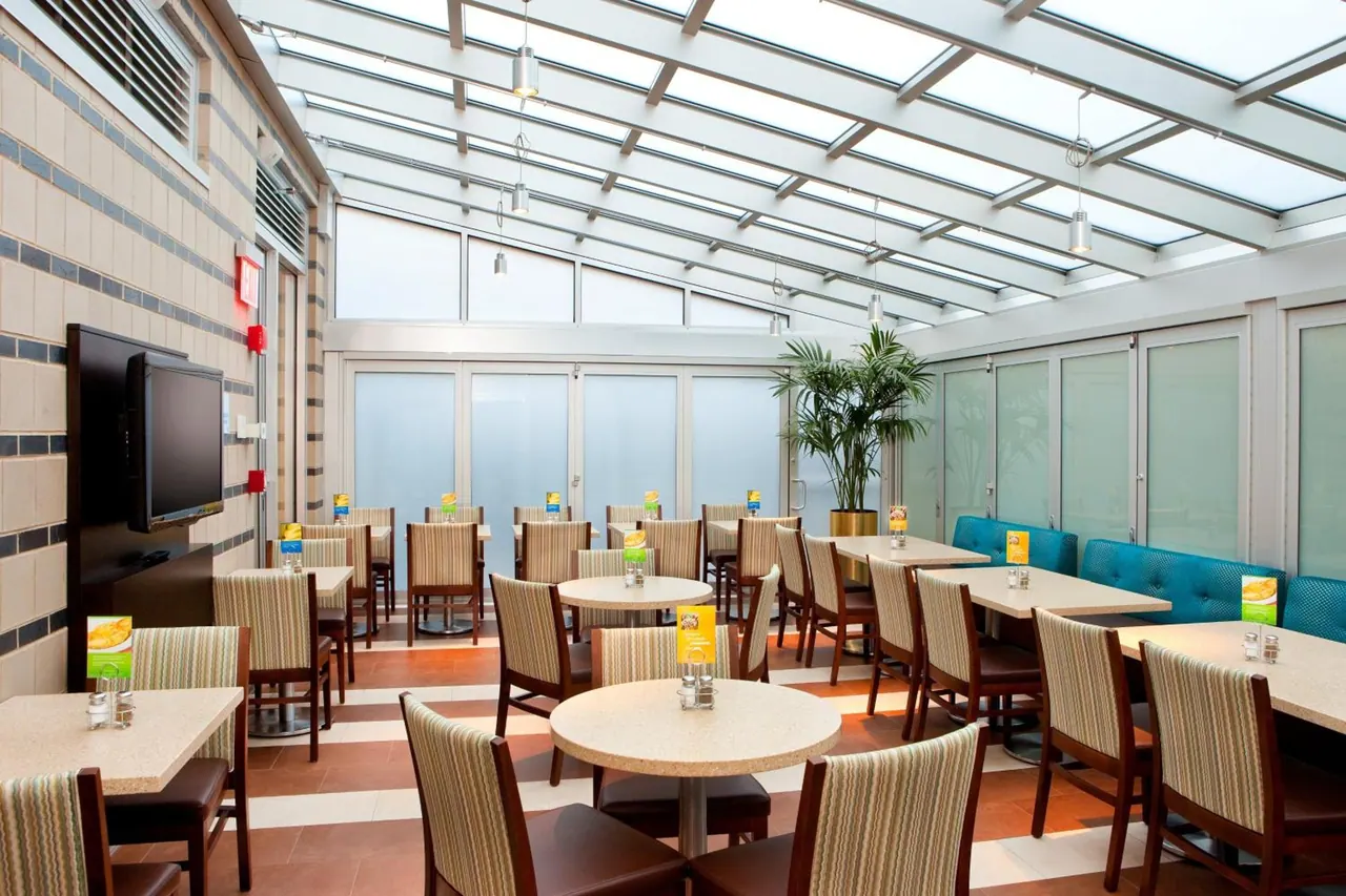 A dining room with a glass roof, rows of tables with striped upholstered chairs, a wall-mounted television, and blue banquette seating along the back wall.