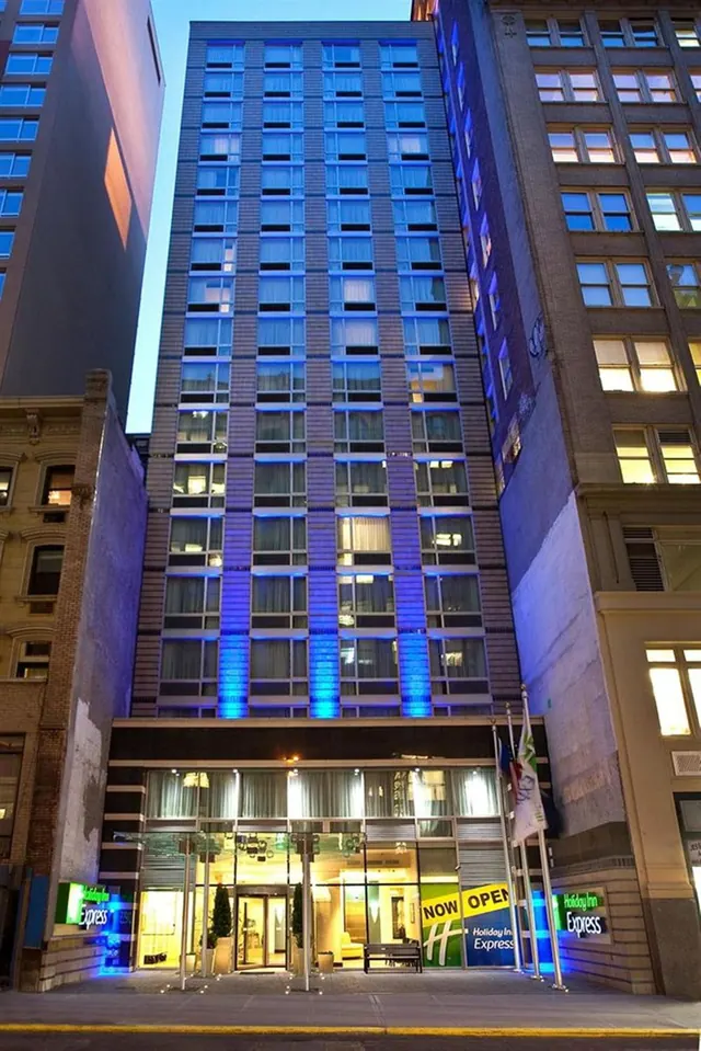 Exterior of a Holiday Inn Express hotel tower lit with blue lights at dusk, with a glass entrance canopy, flags and a Now Open banner at street level.