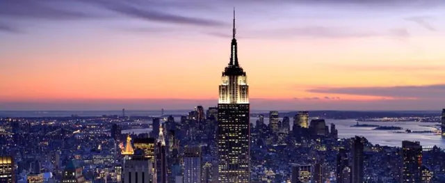 The Empire State Building rises above a city skyline at dusk, with a body of water visible on the right.