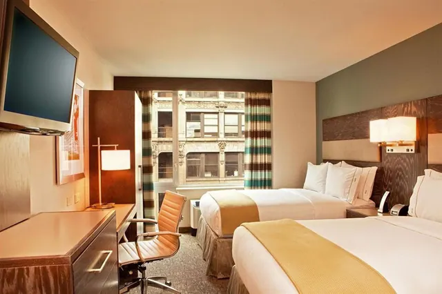 A hotel room with two beds with yellow runners, a wall-mounted television, a desk with a chair, and a window looking out at an ornate building facade.