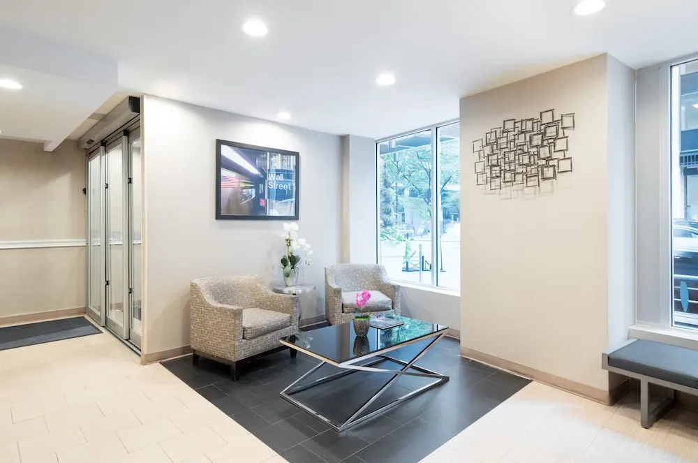 A lobby seating area with two upholstered armchairs, a glass coffee table, a framed Wall Street print, and large windows looking onto a street with trees.