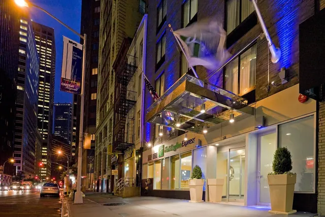 Night view of the Holiday Inn Express entrance on a city street, showing a glass canopy, potted shrubs, and a flag.