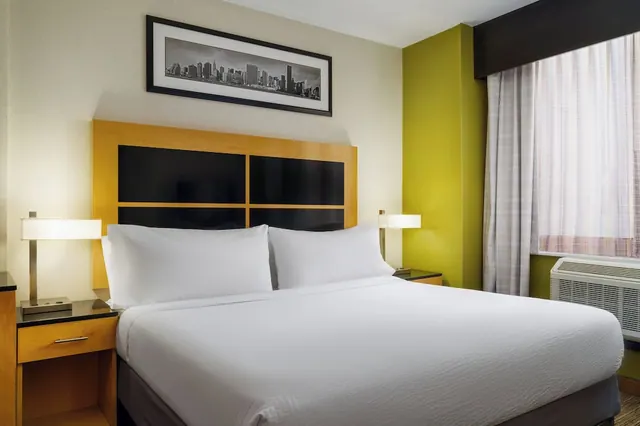 A king bed with white linens sits beneath a framed black-and-white skyline print, with bedside lamps on each side and a window air conditioner unit below the sheer-curtained window.