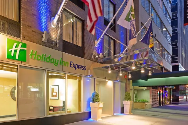 Night exterior of a Holiday Inn Express hotel entrance with illuminated signage, two flags on poles, and potted plants on the sidewalk.