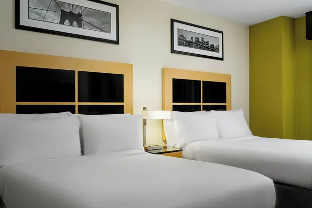 Two double beds with white bedding sit beneath black and white framed prints of city and bridge scenes, with a bedside lamp on a nightstand between them.
