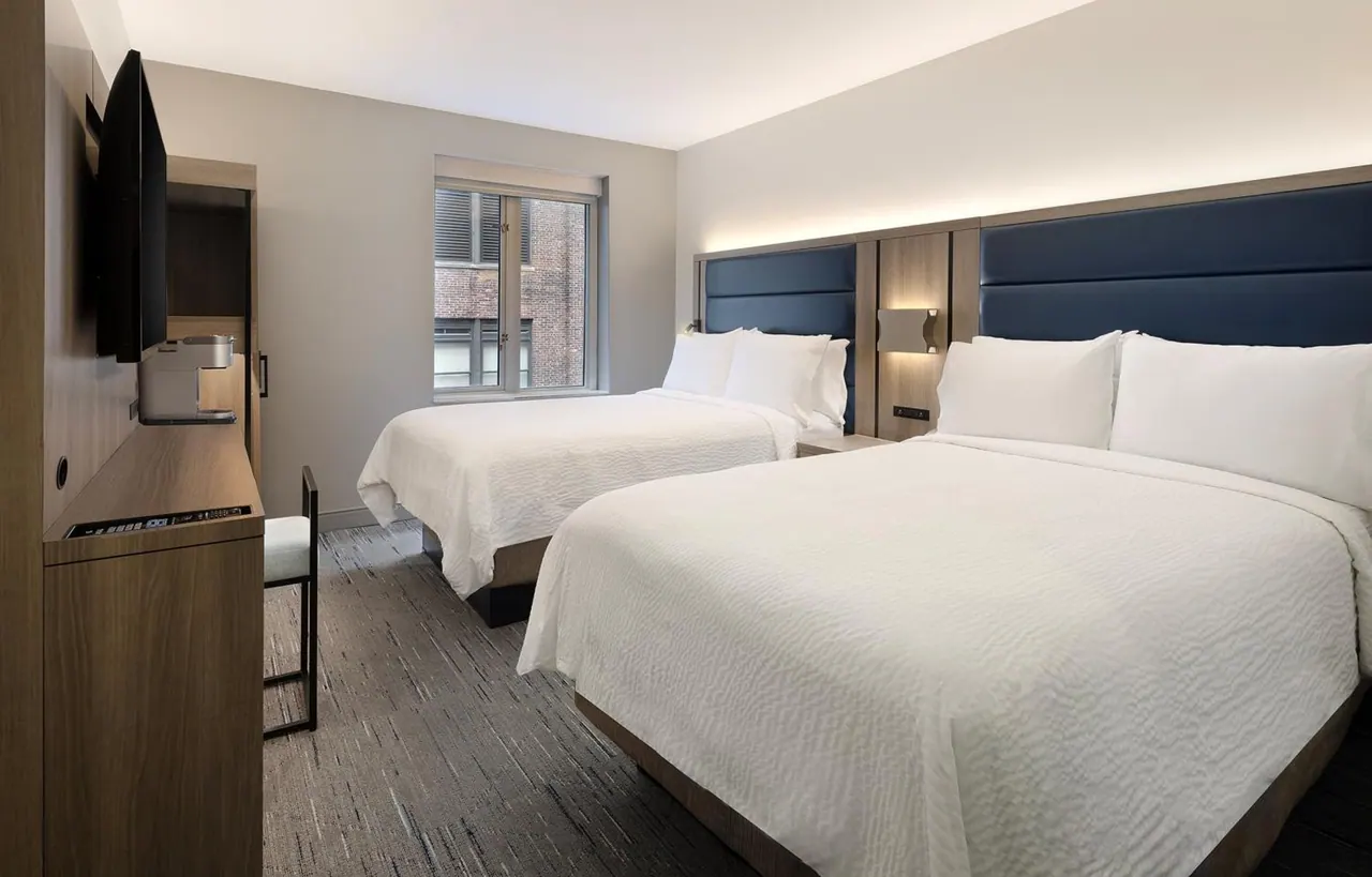 A hotel bedroom with two white-covered beds with navy upholstered headboards, a wall-mounted television, a wood desk with a coffee machine, and a window looking onto a brick building.