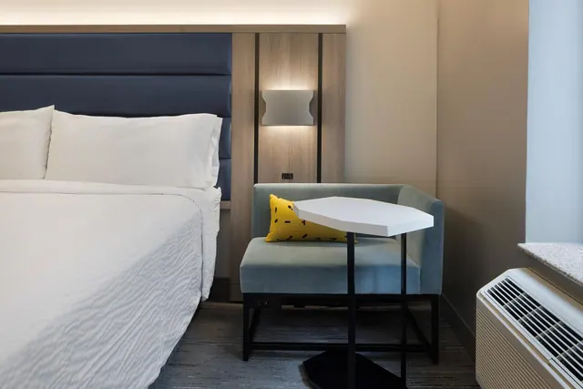 A king bed with white linens sits beside a blue-green armchair with a yellow cushion and a white side table, with a wall-mounted air conditioning unit at the right.