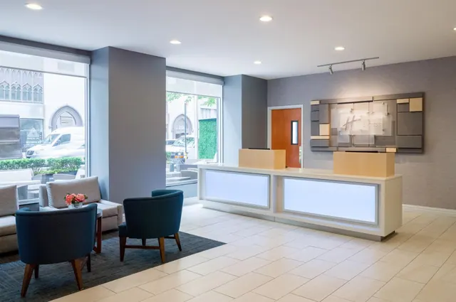 A hotel lobby with a reception desk, lounge seating and large windows looking onto a street with parked vehicles.