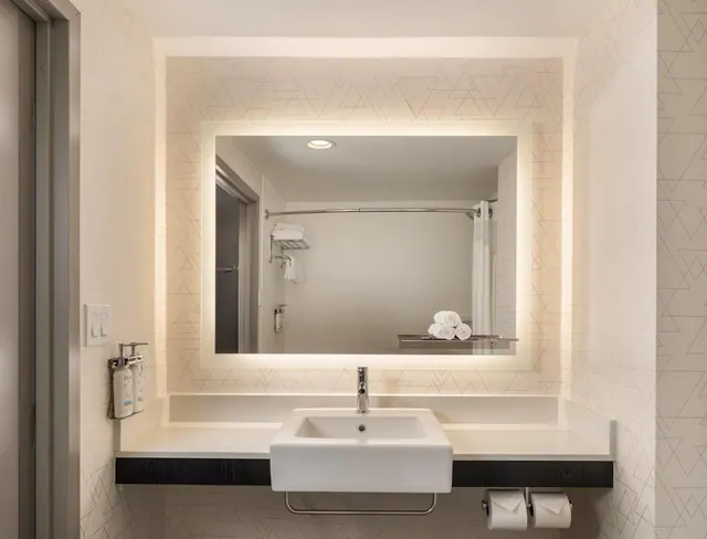 A bathroom vanity with a single white basin, a backlit mirror, wall-mounted toiletry dispensers and toilet roll holders below the counter.