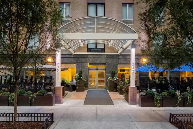 The front entrance of a hotel building under a curved glass canopy, with planters, a glass door and blue umbrellas beside it at dusk.
