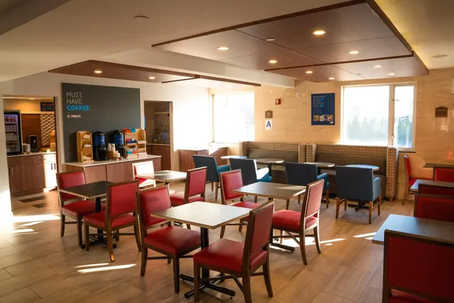 A breakfast dining room with red-upholstered chairs and small tables, a coffee station with dispensers, and booth seating along the windows.