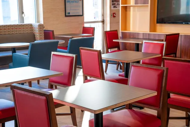 A breakfast dining room with red and blue upholstered chairs, square tables, a booth bench and a wall-mounted television.