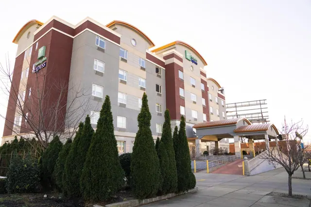 The exterior of a multi-storey Holiday Inn Express building with a covered entrance canopy, a sloped ramp with railings, and tall evergreen shrubs in front.