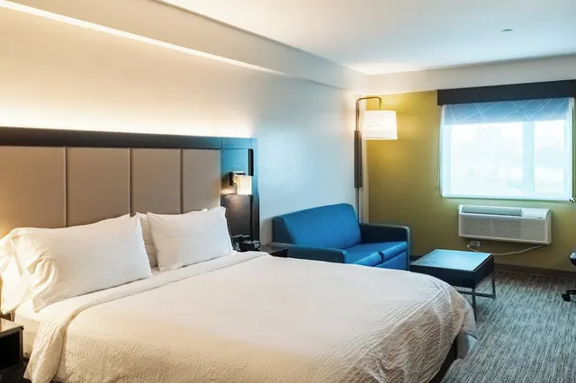 A king bed with white linens sits beside a blue sofa and a window with a wall-mounted air conditioner beneath it.