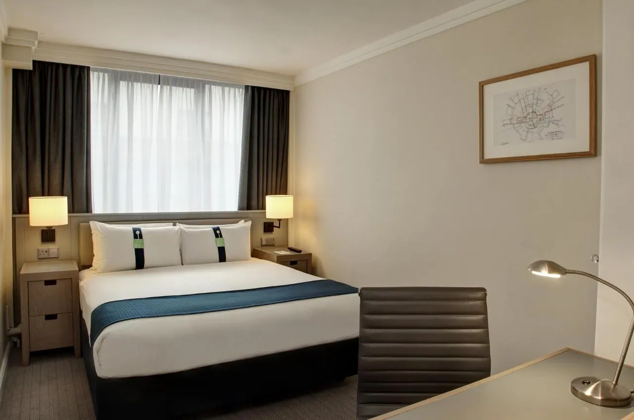 A double bed with a teal runner sits beside a window with sheer curtains, with a framed map on the wall and a desk with a lamp in the foreground.