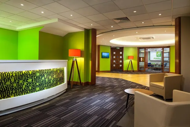 A lobby with a curved white reception desk with an illuminated green front, green walls, armchairs and a round wooden table, with a meeting room visible in the background.