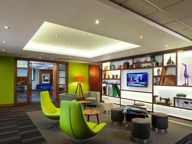 A seating lounge with lime green armchairs, grey stools, small wooden side tables, a wall-mounted television and built-in shelving, with a glass-walled meeting room visible through the doorway.