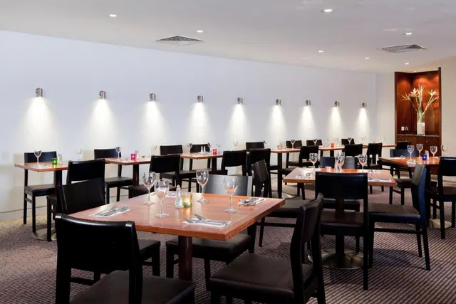 A restaurant dining room with rows of square wooden tables, black leather chairs and spotlights on the white wall.