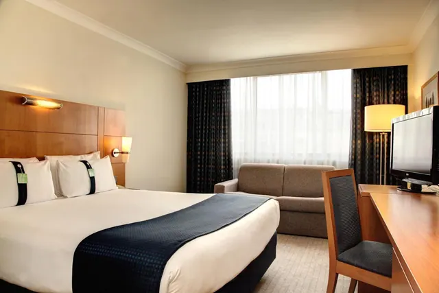 A hotel bedroom with a made double bed, a grey sofa by the window, a floor lamp, and a wooden desk with a flat-screen television.