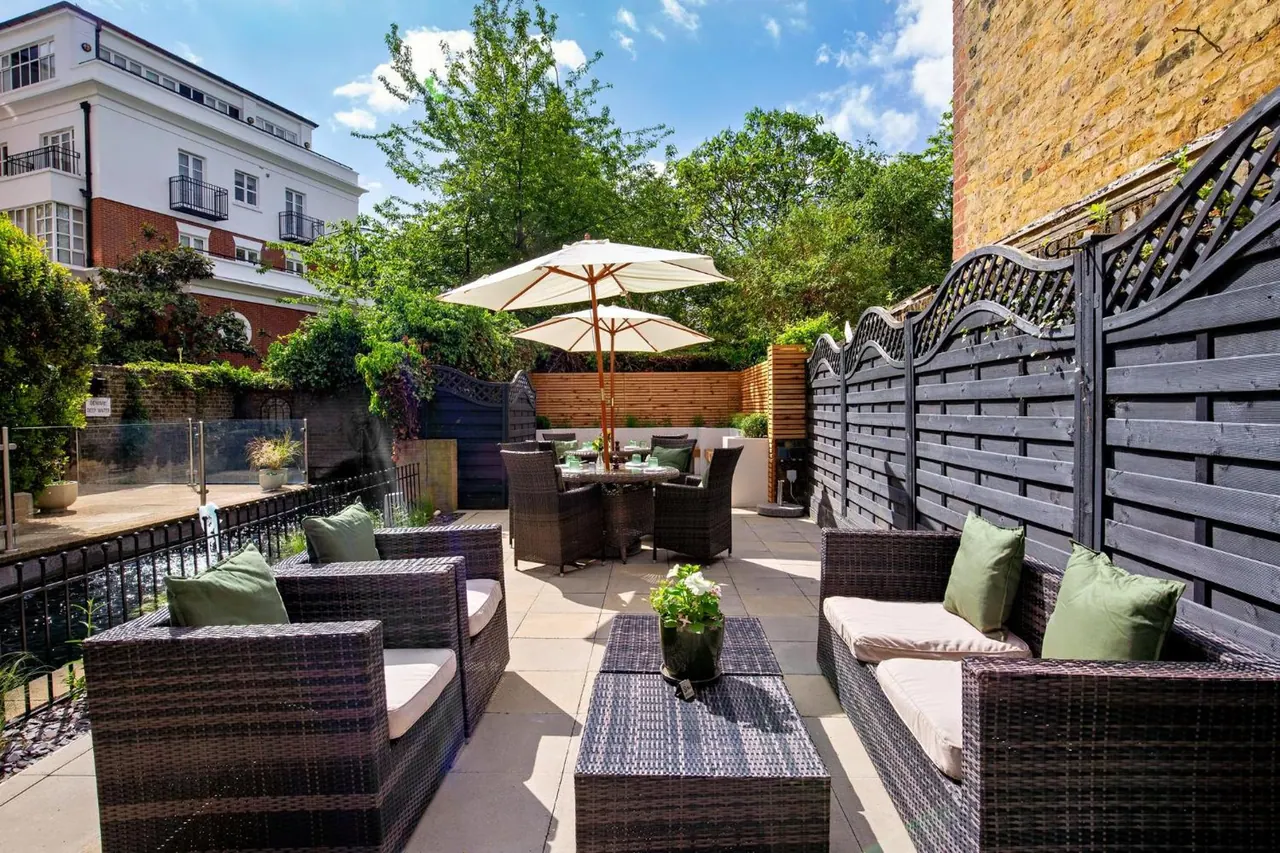 An outdoor terrace with wicker sofas, a dining table under parasols, and a dark wooden fence, with a white building and trees beyond.