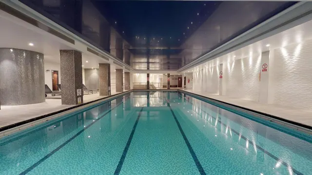 An indoor lap pool with lane lines runs toward a far wall, with sun loungers visible at the far left under a dark reflective ceiling.