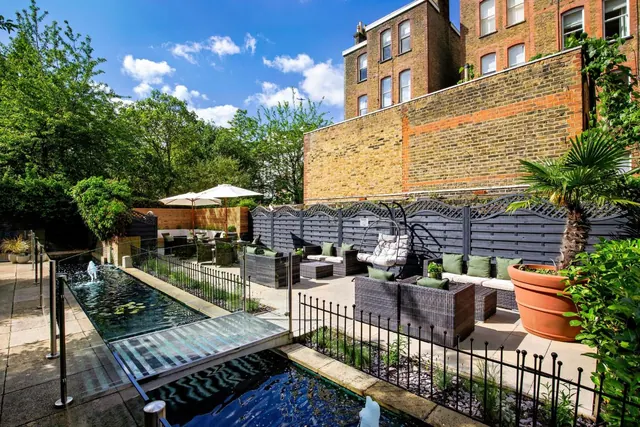 A sunny walled garden courtyard with a narrow water channel, rattan seating with cushions, two parasols and a potted palm, with a brick building behind.