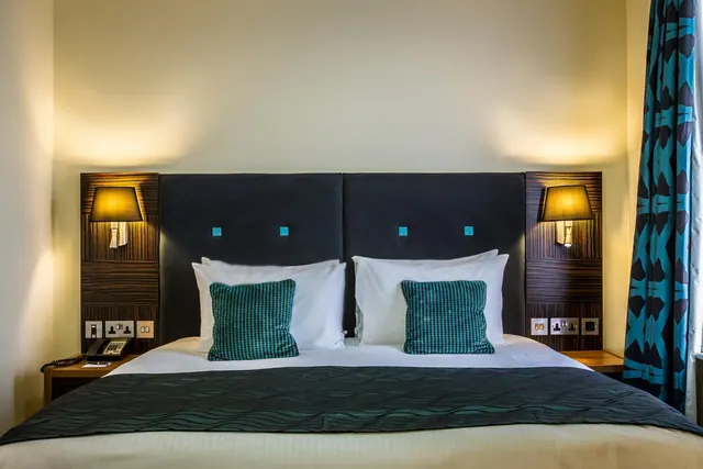 A double bed with white pillows and teal cushions sits beneath a dark upholstered headboard, flanked by two wall lamps and a bedside telephone.