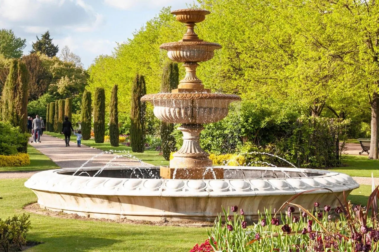 A tiered stone fountain stands in a formal garden beside a gravel path, with cypress trees, tulips in the foreground, and a few people walking in the background.