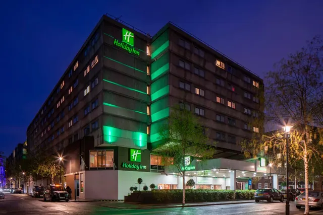 The exterior of a Holiday Inn building at dusk, with illuminated green signage, parked cars and a street lamp in view.