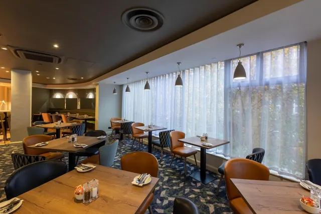 A restaurant dining room with wooden tables, dark upholstered chairs, pendant lights and tall sheer-curtained windows looking onto trees.