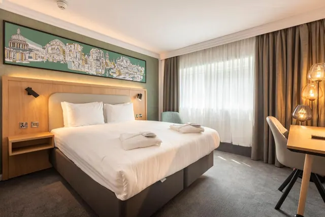 A double bed with white linen and folded towels sits beside a sheer-curtained window, with a framed London skyline illustration on the green wall and a desk on the right.